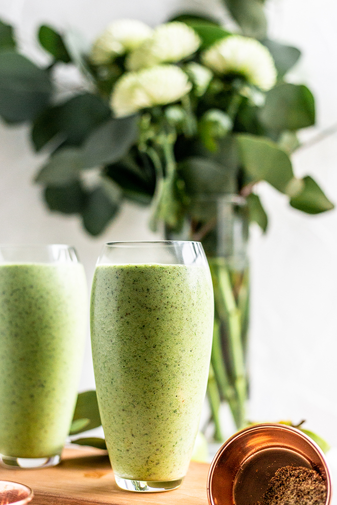 two green apple smoothies with a flower arrangements on a wood board with a white background