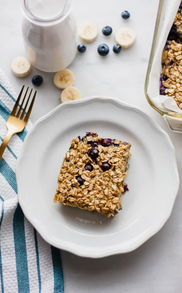 Banana Blueberry Baked Breakfast Oatmeal on a white plate with a milk glass, a dish towel and a gold fork