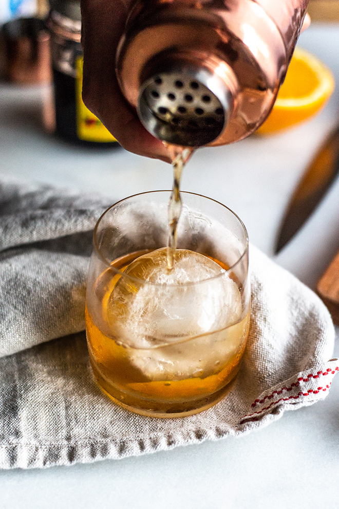 pouring an old fashioned cocktail on a linen napkin with a hand and shaker