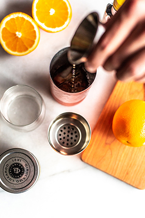 pouring bourbon into a copper shaker on a shite background with oranges, and a wood cutting board