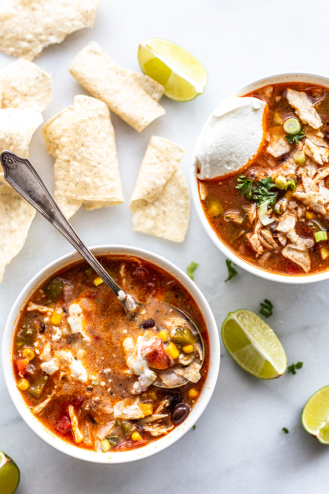 red tortilla soup with Greek yogurt that has been mixed with another soup on the size, limes, chips all shot on a white marble background 