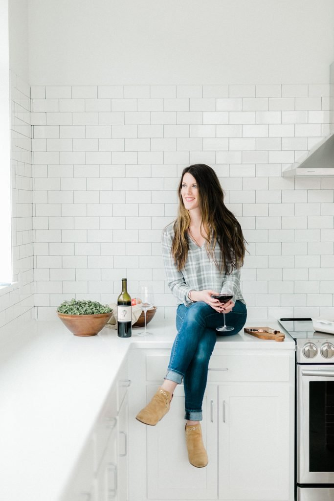 girl in white kitchen in a plaid shirt with brown hair and a wine glass