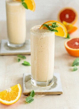 dairy free orange creamsicle milkshake in a slender glass with a coaster on a cream backdrop, mint and oranges