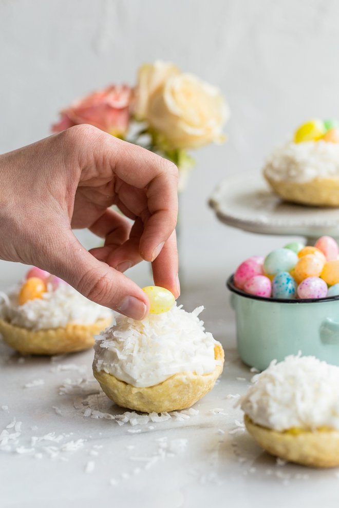 easy mini coconut cream pies with pastel jelly beans on marble with coconut on top and roses and a jelly bean bowl in the background with a hand placing a yellow jellybean