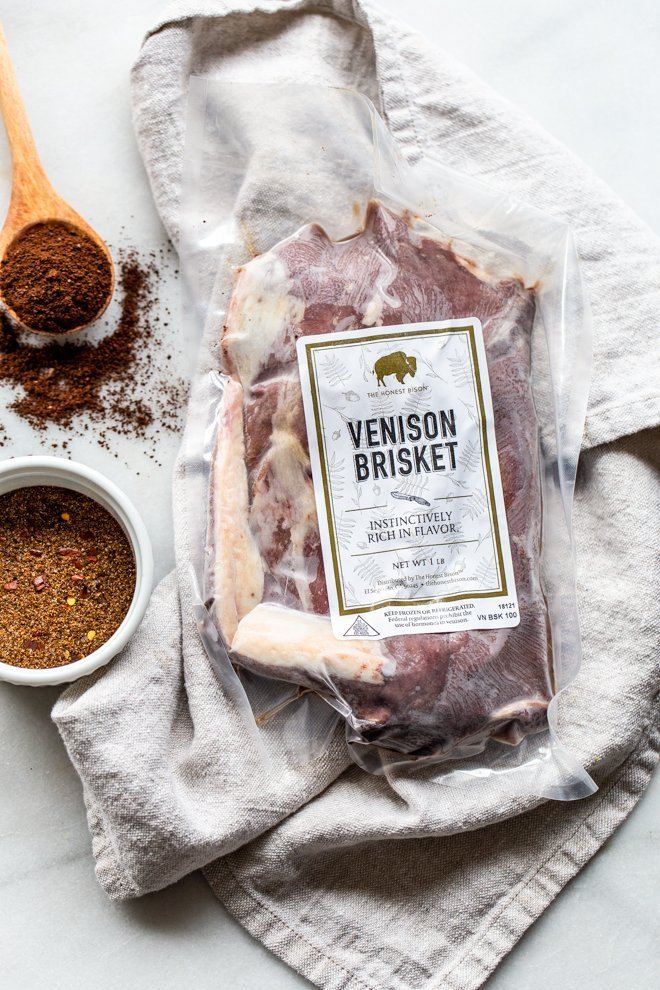 venison brisket package on a cream linen with a bowl of rub and a wooden spoon of coffee