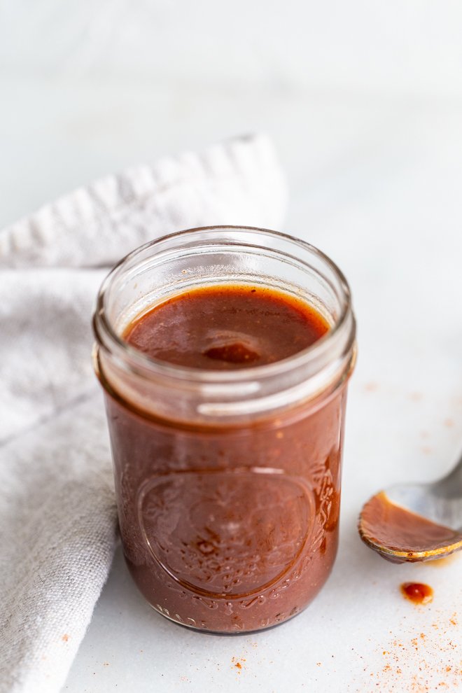 paleo BBQ sauce in a glass jar with a cream linen on a white background with a silver spoon