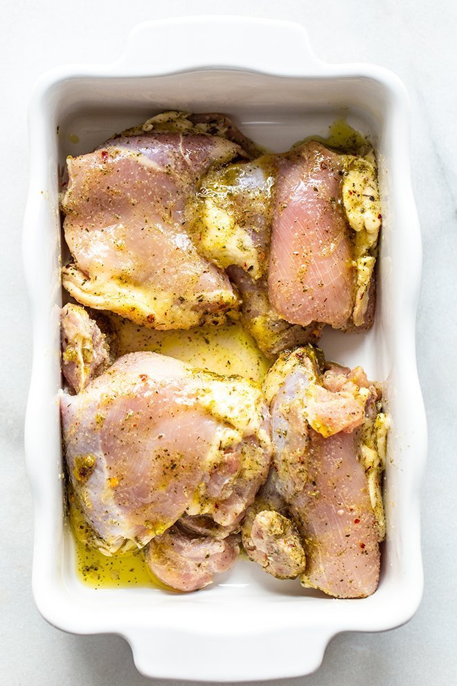 raw chicken thighs marinating in a white casserole dish with olive oil and spices