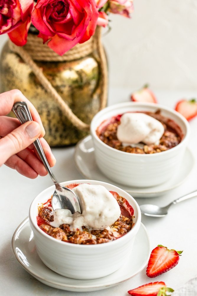 Gluten Free Strawberry Crisp for Two on a white background with strawberries on a white background with vanilla ice cream and a hand scooping in