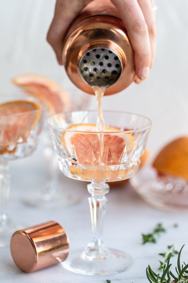 pouring a grapefruit cocktail with a grapefruit slice, a shaker cap, herbs on a white background