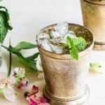 a mint julep cocktail with a glass of mint and flowers on a cream background