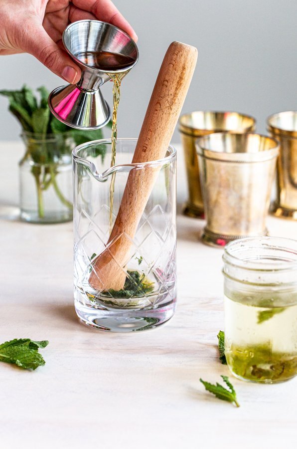 muddling a mint julep cocktail in a glass pitcher on a cream background