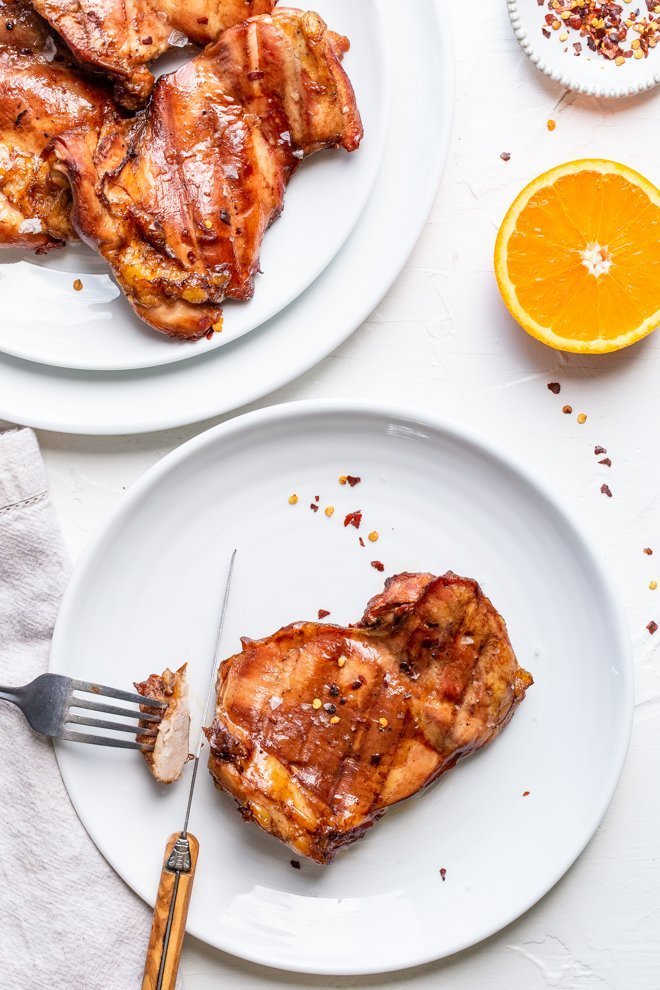 a smoked chicken thigh on a plate on a white background. An orange half, red pepper flakes and a platter of chicken thighs are visible