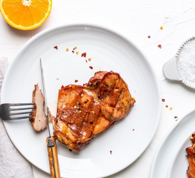 a chicken thigh on a plate on a white background. An orange half, red pepper flakes and a platter of chicken thighs are visible