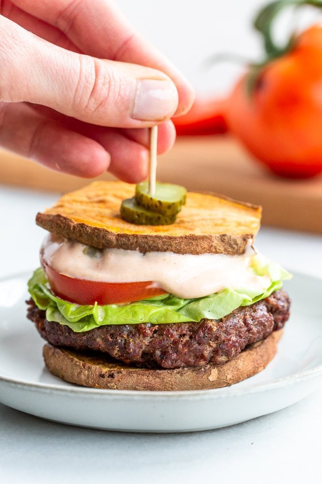 a burger with special sauce on a white plate with a hand placing a toothpick with pickles