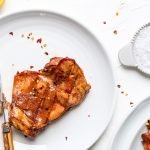 a smoked chicken thigh on a plate on a white background. An orange half, red pepper flakes and a platter of chicken thighs are visible