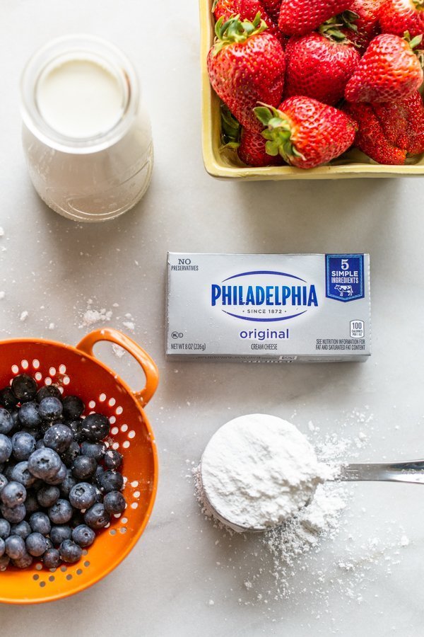 blueberries, strawberries, cream cheese, powdered sugar, and cream on a white board. The ingredients for the easiest berry fool recipe. 