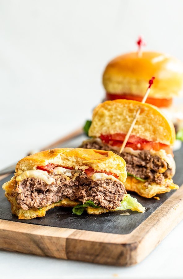 juicy elk burgers with lettuce, tomato and cheese with a bite out of one on a wood and slate board with a white background