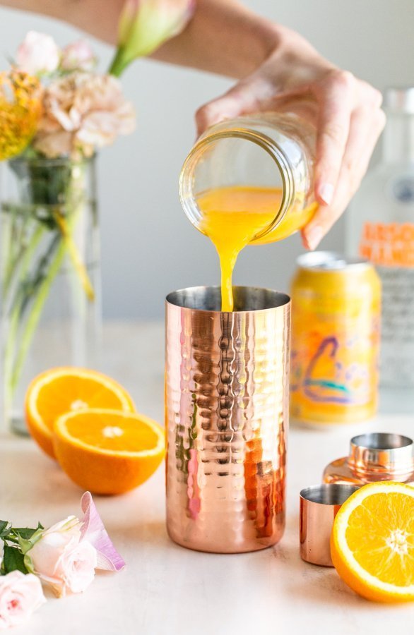 pouring orange juice into a copper cocktail shaker with oranges in the background