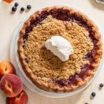 peach blueberry pie with a crumble topping on a cream board with a striped linen napkin