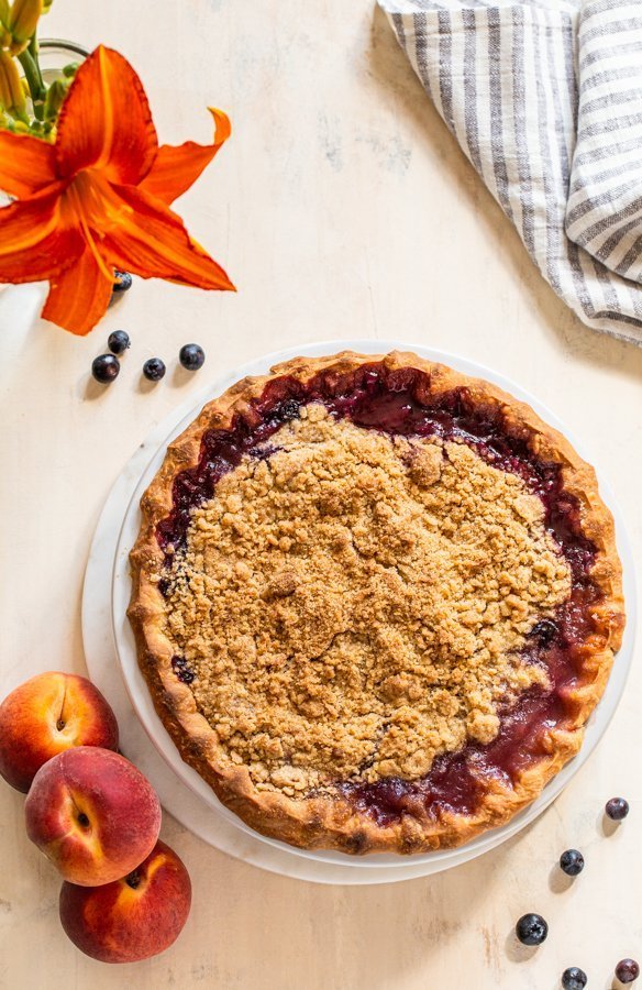 peach blueberry pie with a crumble topping on a cream board with a striped linen napkin