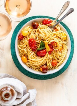 summer pasta with tomatoes and cheese in a white bowl on a blue charger with rosé wine and a fork and a spoon twirling the pasta