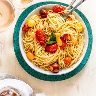 summer pasta with tomatoes and cheese in a white bowl on a blue charger with rosé wine and a fork and a spoon twirling the pasta