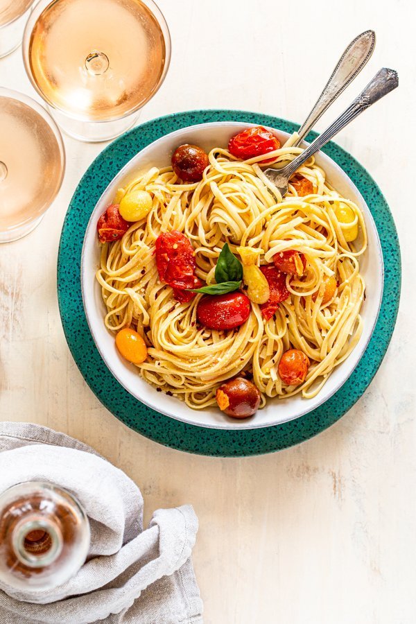 summer pasta with tomatoes and cheese in a white bowl on a blue charger with rosé wine and  a fork and a spoon twirling the pasta