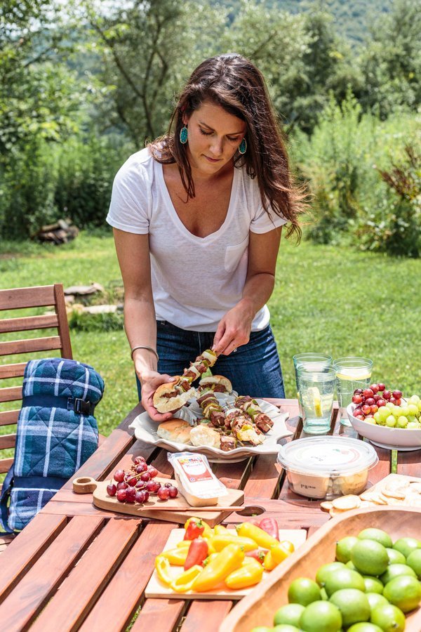 woman with brown hair and white shirt serving a kabob on a table spread with tailgate food. 