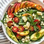 a late summer salad with corn, tomatoes and peaches in a white bowl with avocado slices and green dressing