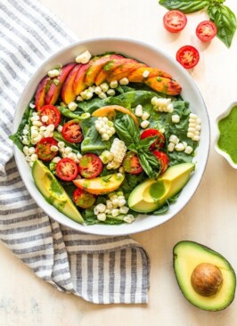 a late summer salad with corn, tomatoes and peaches in a white bowl with avocado slices and green dressing