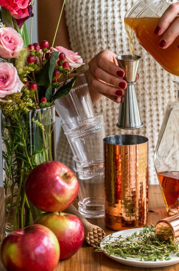 pouring cider syrup into a copper cocktail shaker with stacked glasses, flowers and apples