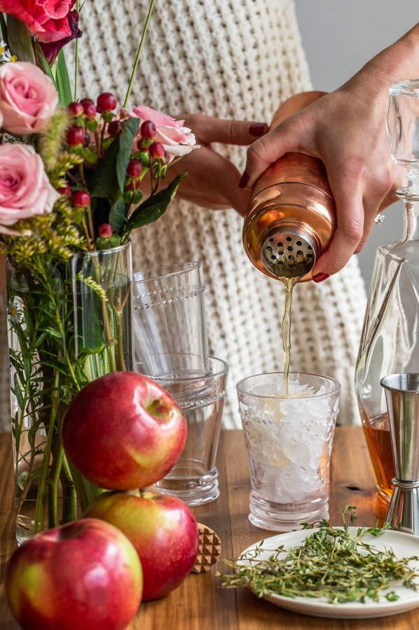 pouring cider bourbon smash from a copper cocktail shaker into a glass with stacked glasses, flowers and apples