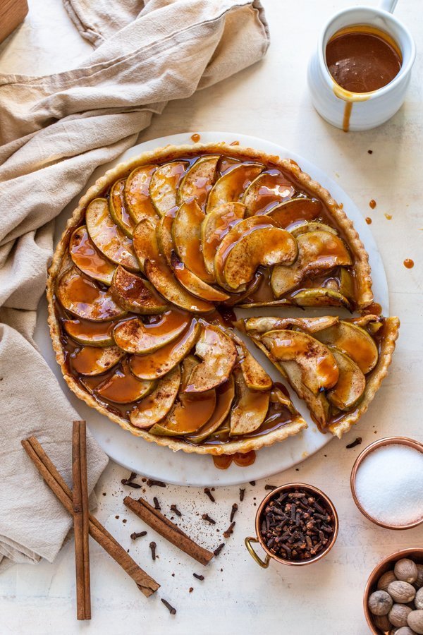 caramel apple tart with a slice cut out of it with cinnamon, cloves and nutmeg