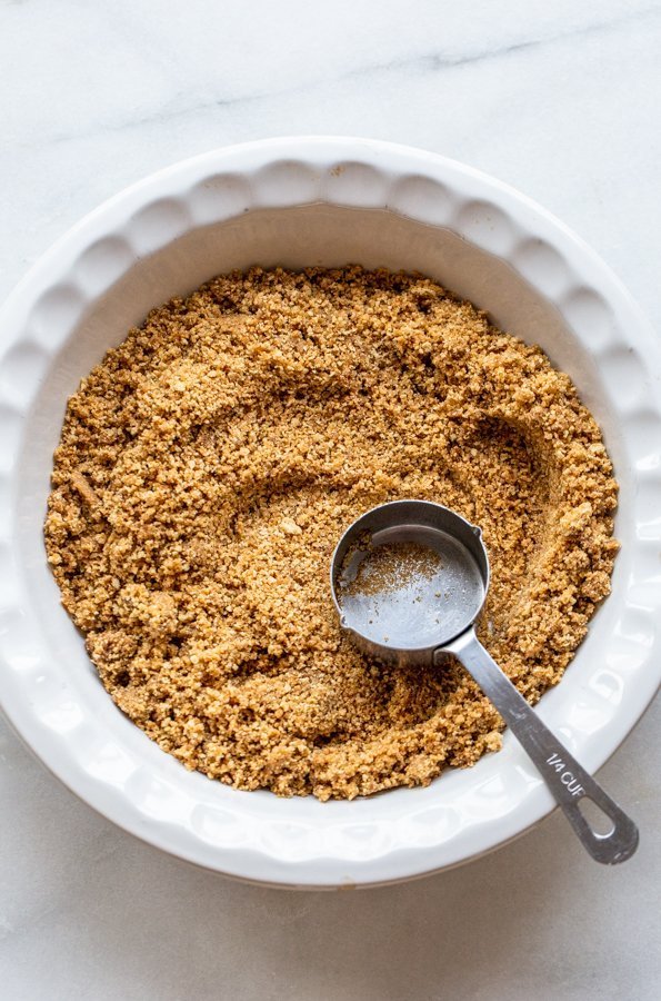 graham cracker crust in a white pie plate being pressed out with a measuring cup