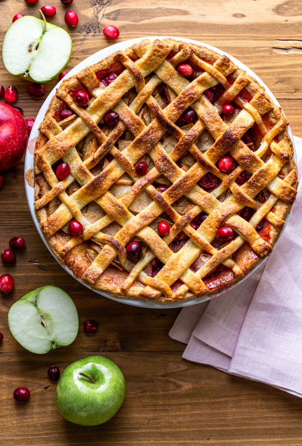 apple cranberry pie on a wood board with a pink napkin, apples and cranberries