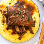 venison short ribs on a light blue plate with polenta. Knife, fork and a blue stripe linen