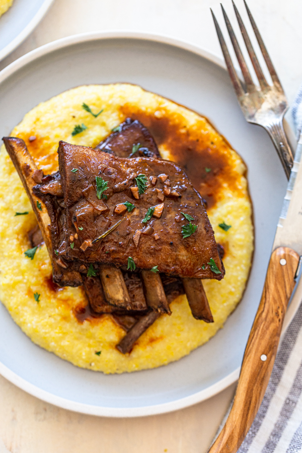 venison short ribs on a light blue plate with polenta. Knife, fork and a blue stripe linen