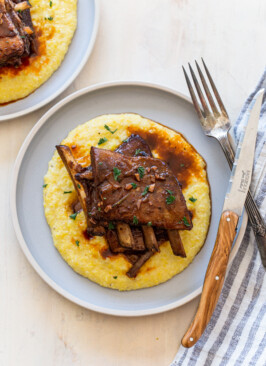 venison short ribs on a light blue plate with polenta. Knife, fork and a blue stripe linen