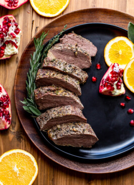 sliced bison tenderloin on a black plate with oranges and pomegranates on a wood table