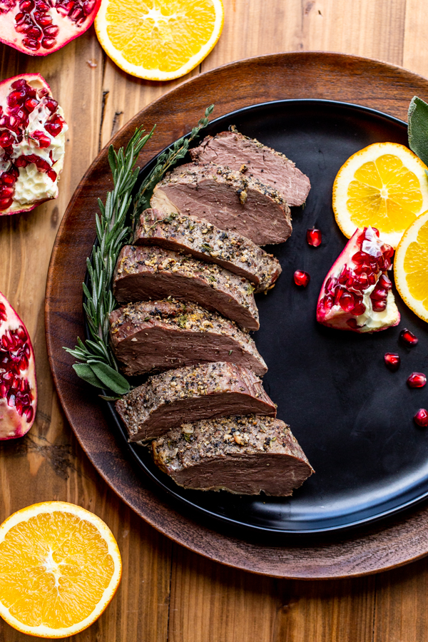 sliced bison tenderloin on a black plate with oranges and pomegranates on a wood table