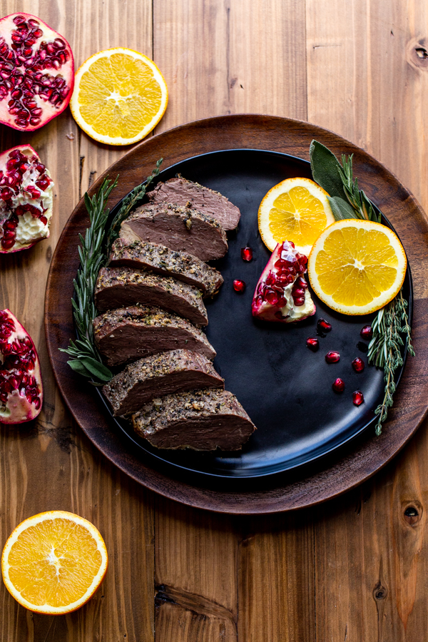 sliced bison tenderloin on a black plate with oranges and pomegranates on a wood table