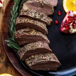 sliced bison meat on a black plate with oranges and pomegranates on a wood table