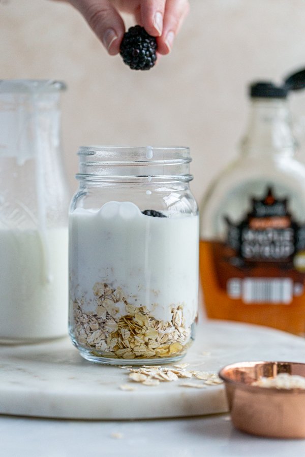 a blackberry being dropped into a jar of overnight oats