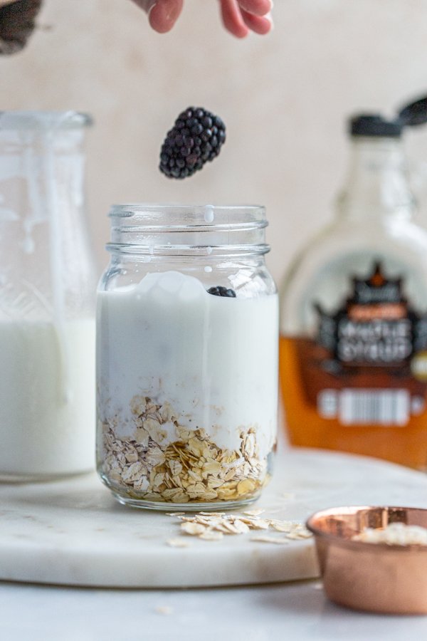 a blackberry being dropped into a jar of overnight oats 