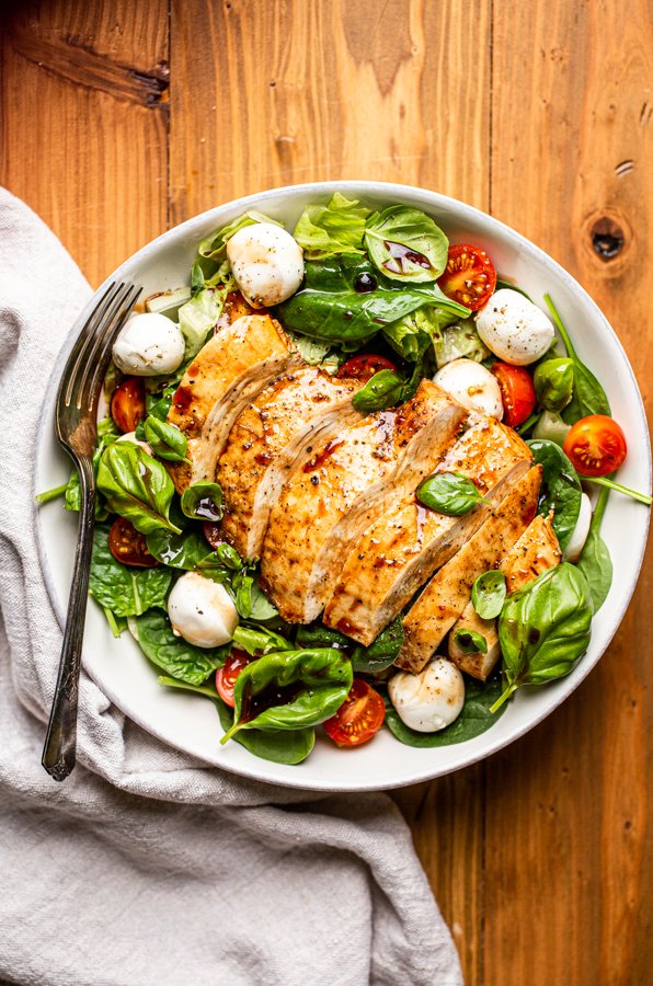 Caprese chicken salad in a white bowl on a wood table with a napkin and fork