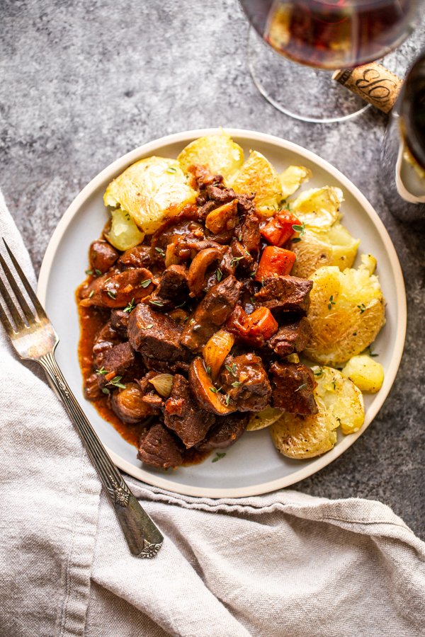 venison bourguignon on a plate with smashed potatoes. Wine in the background.