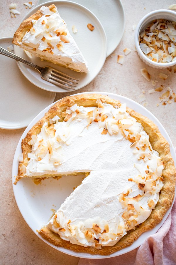 coconut cream pie on a beige counter with toasted coconut and a pink napkin and a slice cut out