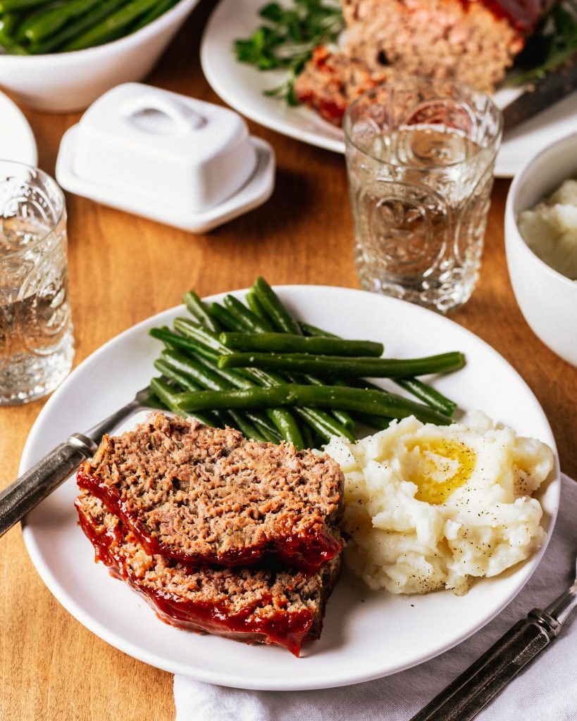 2 slices of venison meatloaf on a white plate with green beans, and mashed potatoes topped with butter