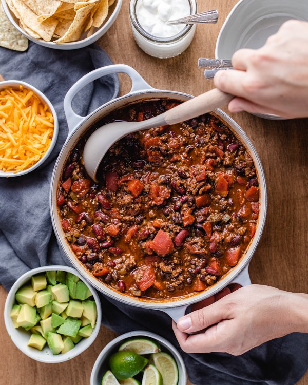 venison chili in a large pot with a ladle on a wood table with toppings spread all around it