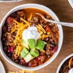 Venison chili, above shot, in a bowl topped with shredded cheddar cheese, chopped avocado and a dollop of sour cream